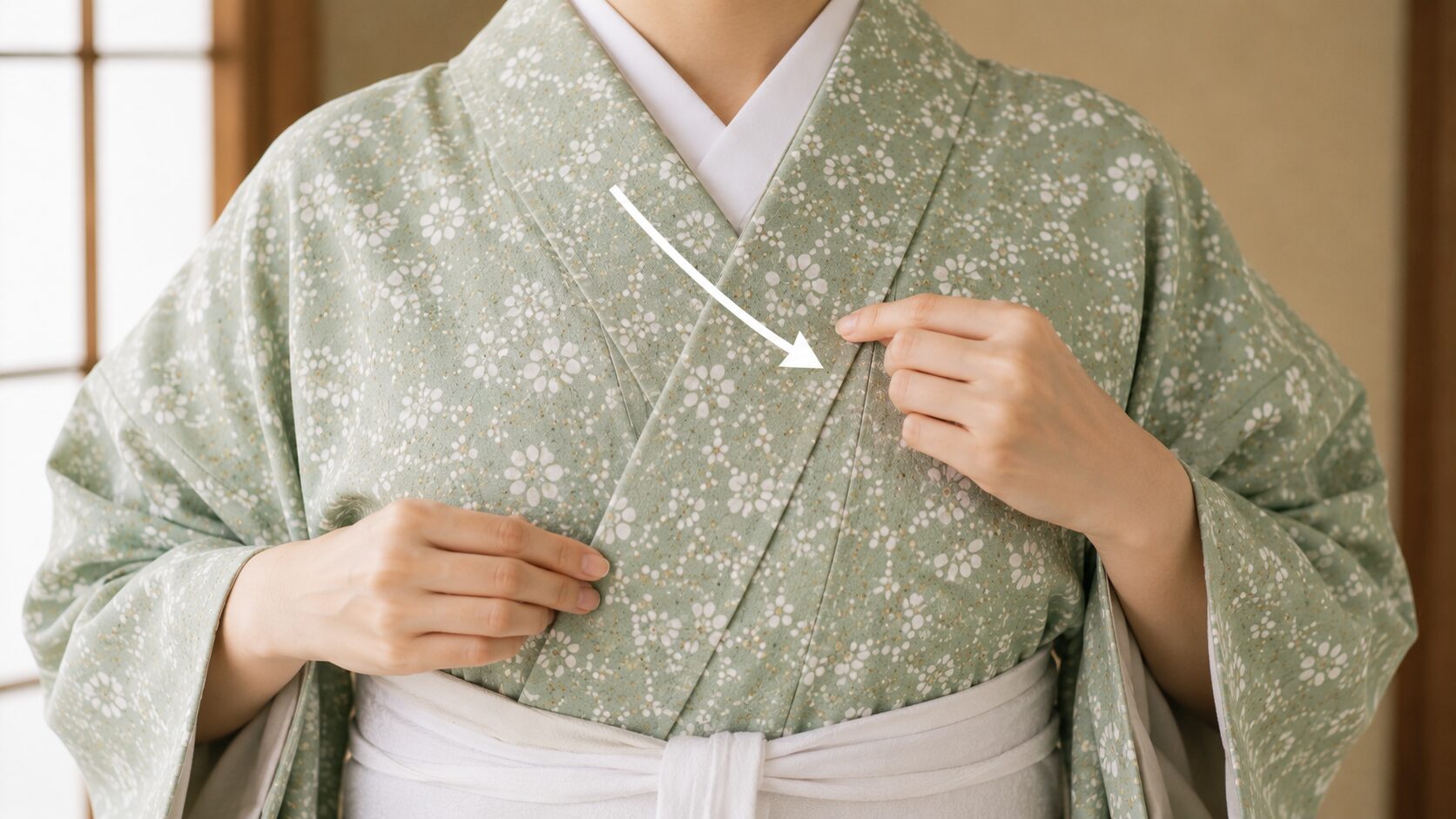 Hands wrapping a celadon green floral kimono left over right across the chest, with an arrow showing the correct collar direction