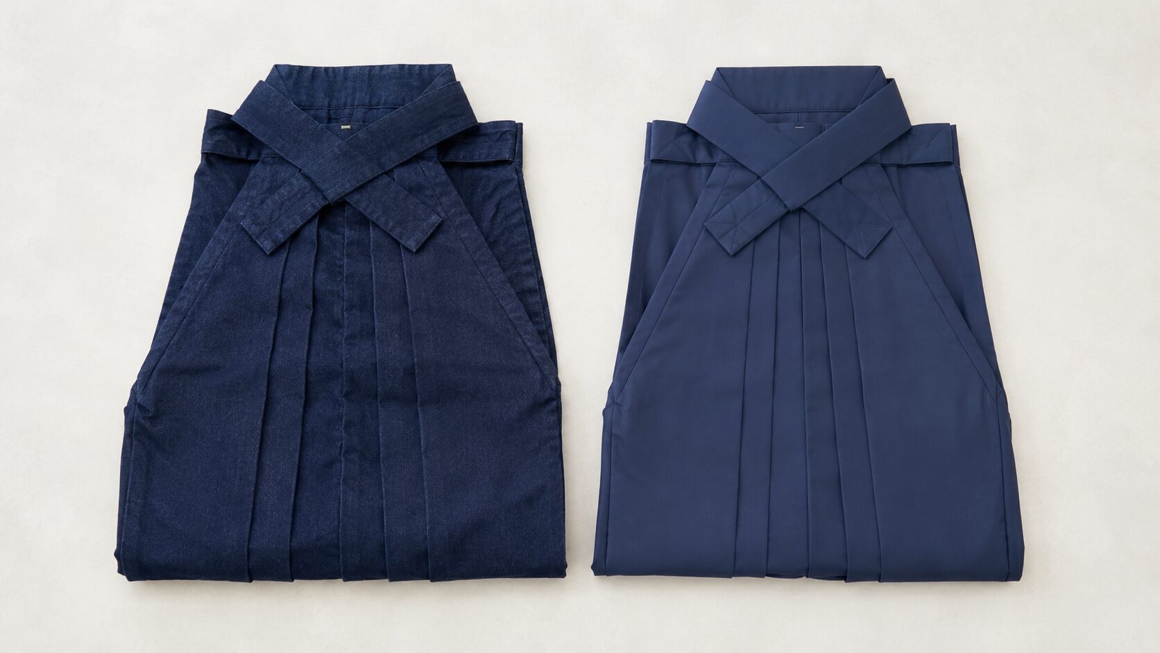 Indigo-dyed cotton hakama and lighter synthetic hakama folded side by side, showing the difference in fabric texture and pleat structure
